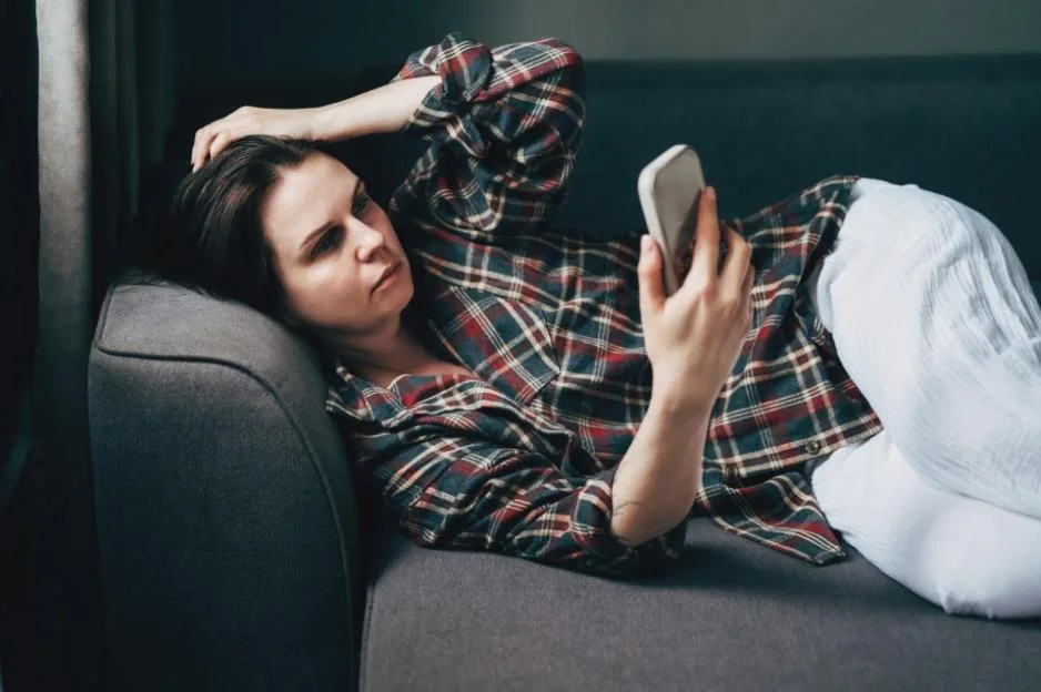 A stressed woman lies on a sofa at home, holding her head in one hand and looking anxiously at her smartphone.