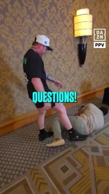 A man wearing a black t-shirt and white hat stands over another man lying on the floor in a hotel hallway.