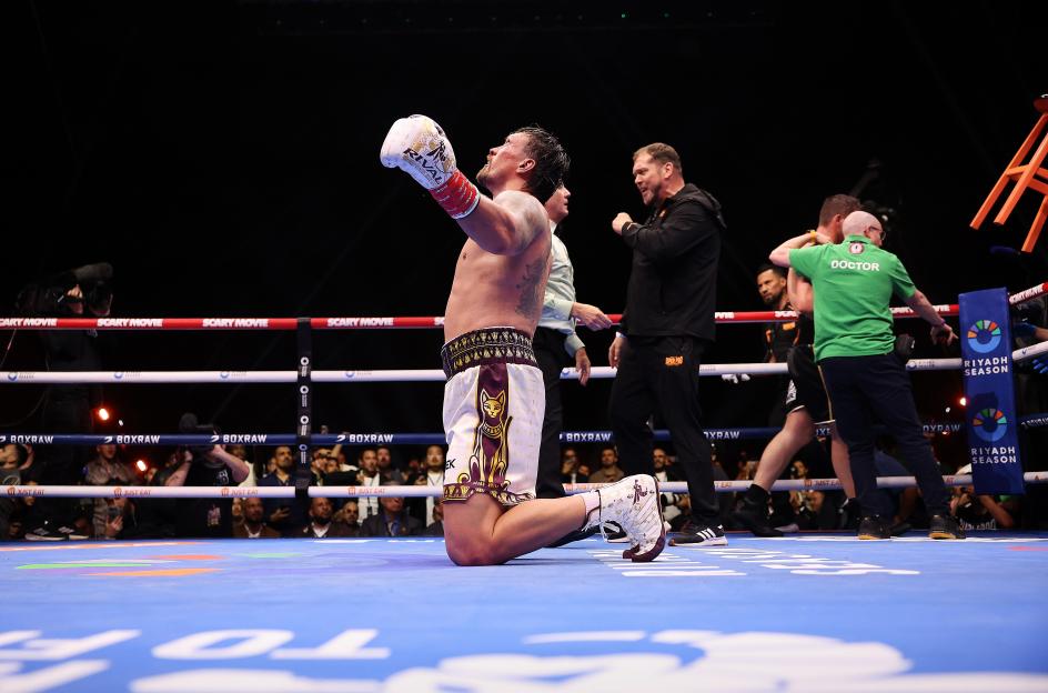 A boxer kneels in the ring with his arm raised in victory, surrounded by his team and a doctor, with the audience in the background.