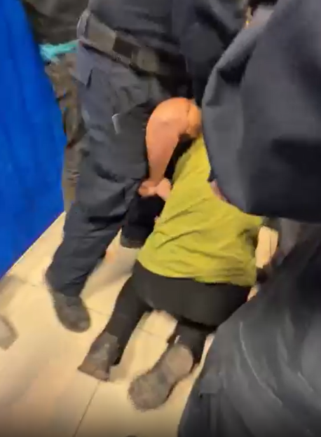 A person in a lime green shirt and black pants is being held down on the floor by two men in dark uniforms.