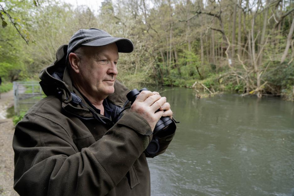Man wearing a cap and jacket, holding binoculars while looking over a river in a wooded area.