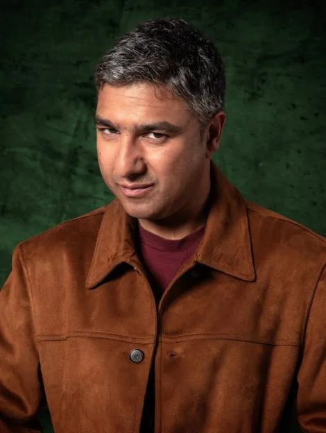 Sanjeev Bhaskar, framed in gold, on a patterned wall with lit candles, a skull, quill, and bird statue on a mantelpiece.