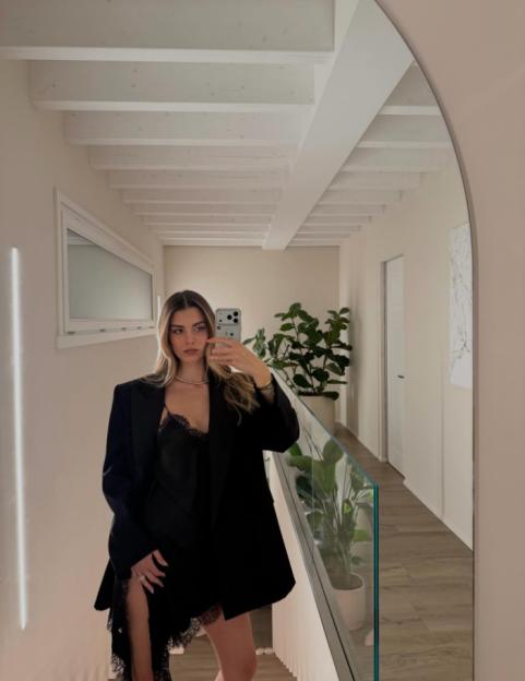 A woman in a black blazer and dress with lace trim takes a mirror selfie.