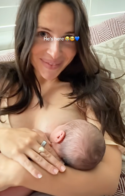 A woman holding a newborn baby to her chest, with "He's here" and crying/blue heart emojis across her forehead.
