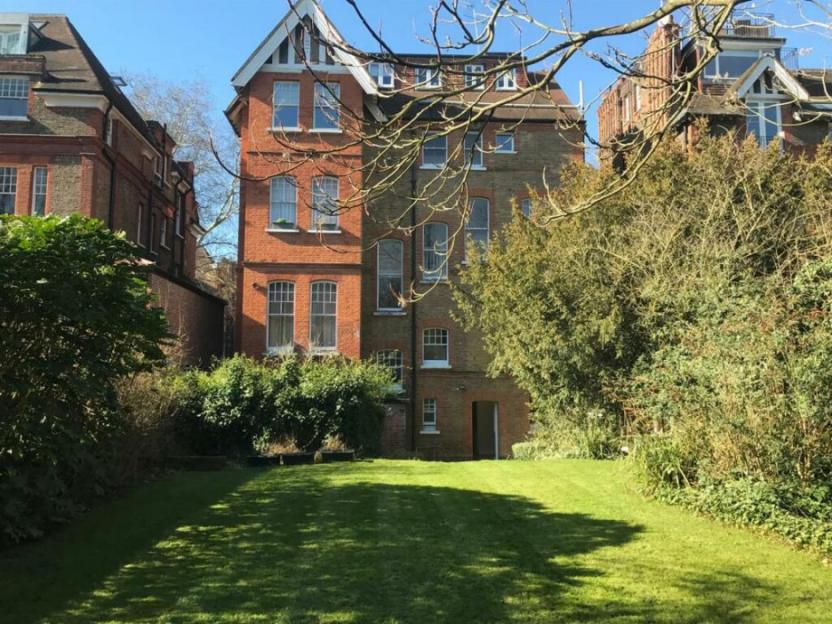 A large, well-maintained lawn with two multi-story brick buildings behind it, surrounded by trees and bushes under a clear sky.