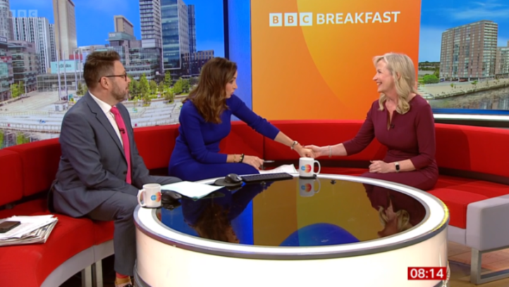 Three people on a TV show set for "BBC Breakfast," with two women shaking hands over a table.