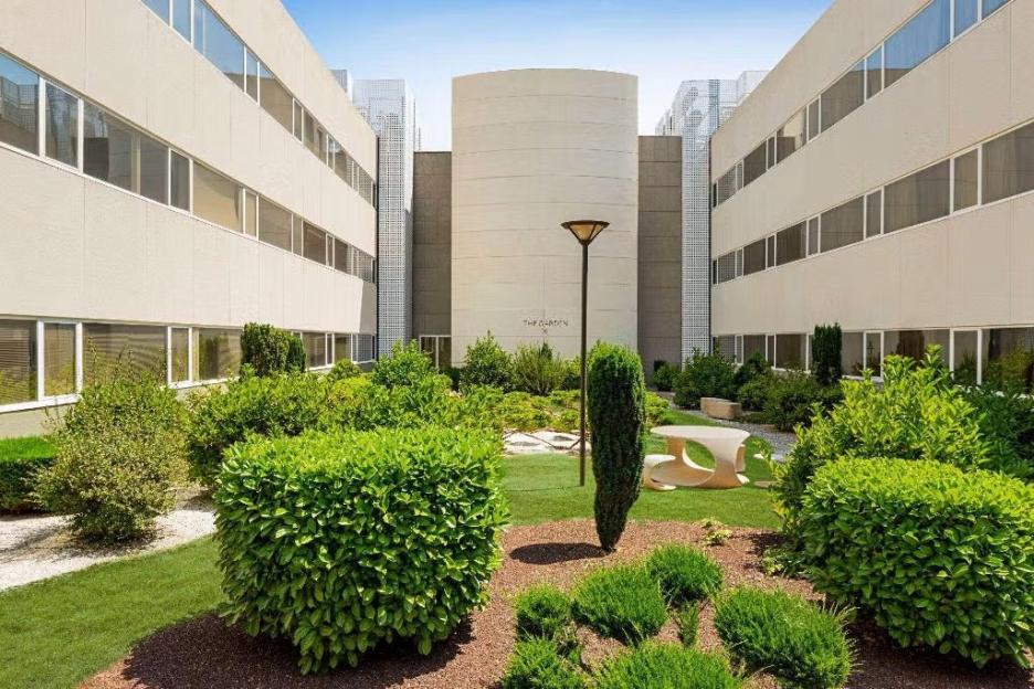 Courtyard garden with manicured shrubs and lawn, flanked by modern buildings, with a sign for "The Garden" on a cylindrical structure.
