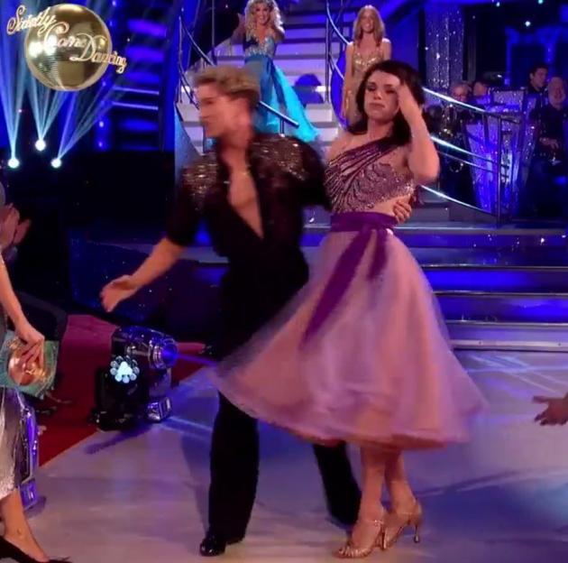A woman in a lilac dress with a purple belt being helped up by a man in a black and sparkly outfit on a dance floor.