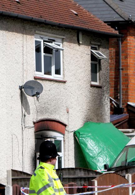 A police officer stands in front of a house damaged by fire, with a green tarp covering part of the scene.
