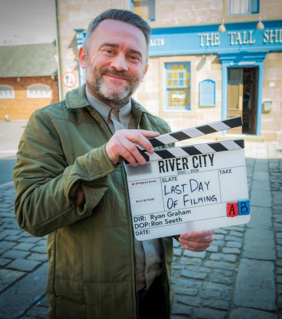 A man in a green jacket holding a clapboard that reads "River City 2002-2026, Last Day Of Filming".
