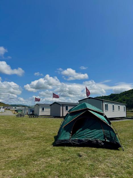 A green tent pitched on a grassy field in front of several caravans under a blue sky with white clouds.