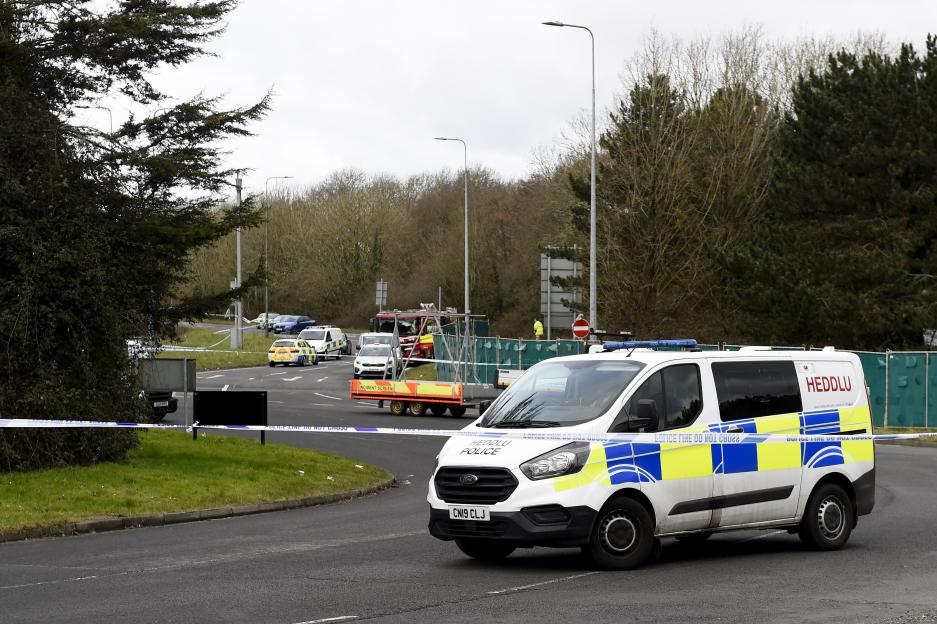 A police vehicle with flashing blue lights and blue and yellow checkered markings blocks a road, marked by white and blue police tape, with other emergency vehicles in the background.
