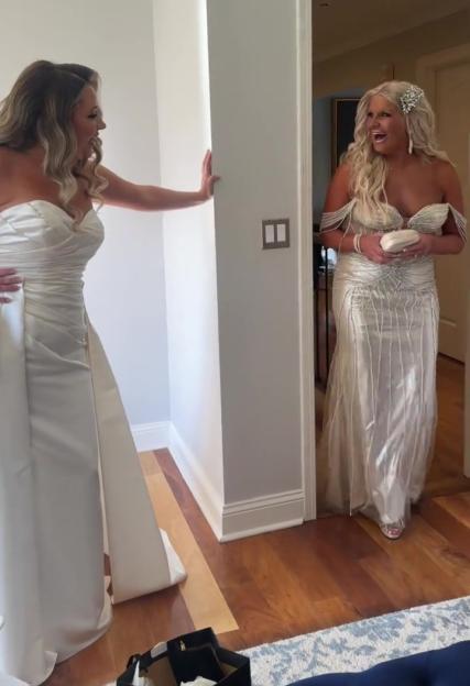 A bride in a white gown and a woman in a shimmering white dress with a decorative hair clip stand opposite each other in a doorway, both with excited expressions.