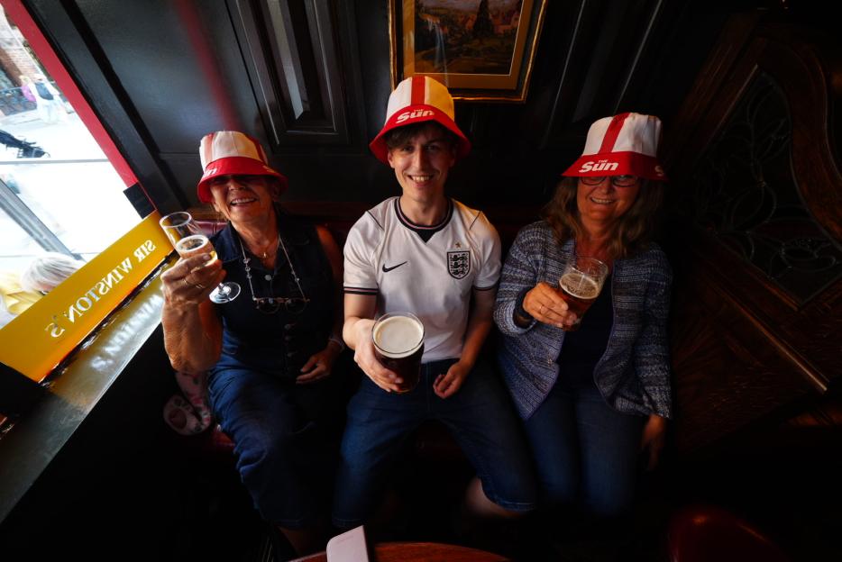 Three people, wearing red and white 'Sun' bucket hats, toast with drinks in a pub.