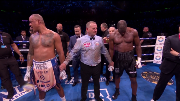 Two boxers and a referee, who is covered in blood, stand in a boxing ring.