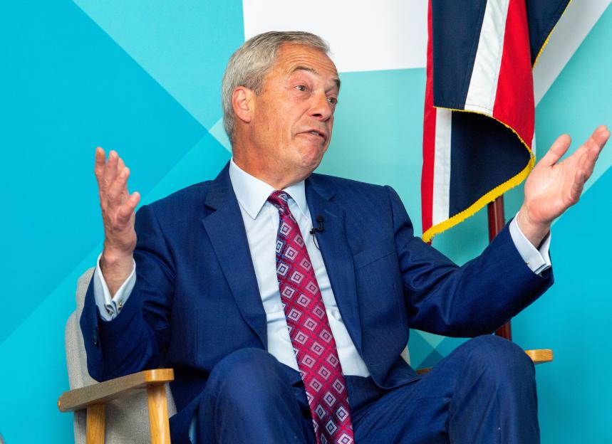 Nigel Farage, leader of the Reform Party, being interviewed while sitting in a chair, gesturing with both hands.