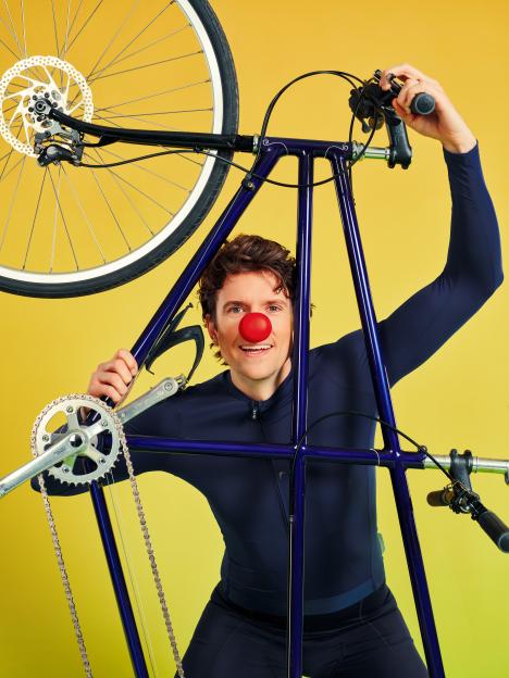 Radio 1 presenter Greg James with a red nose, holding a bicycle above his head.