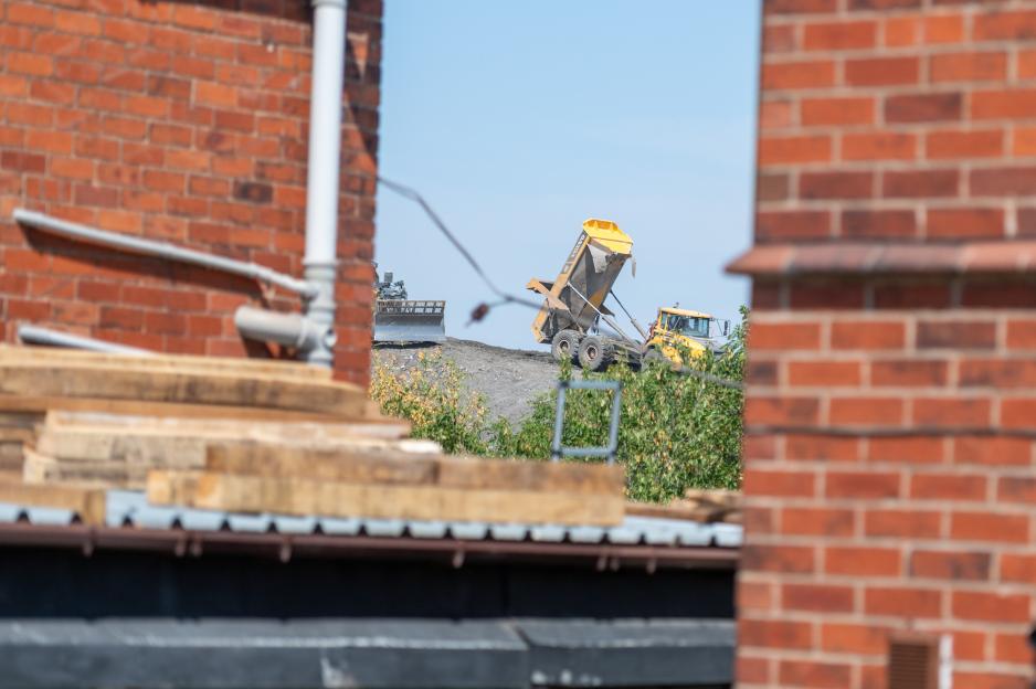 A yellow dump truck with its bed raised, dumping material at a landfill site, seen through an opening between brick walls.