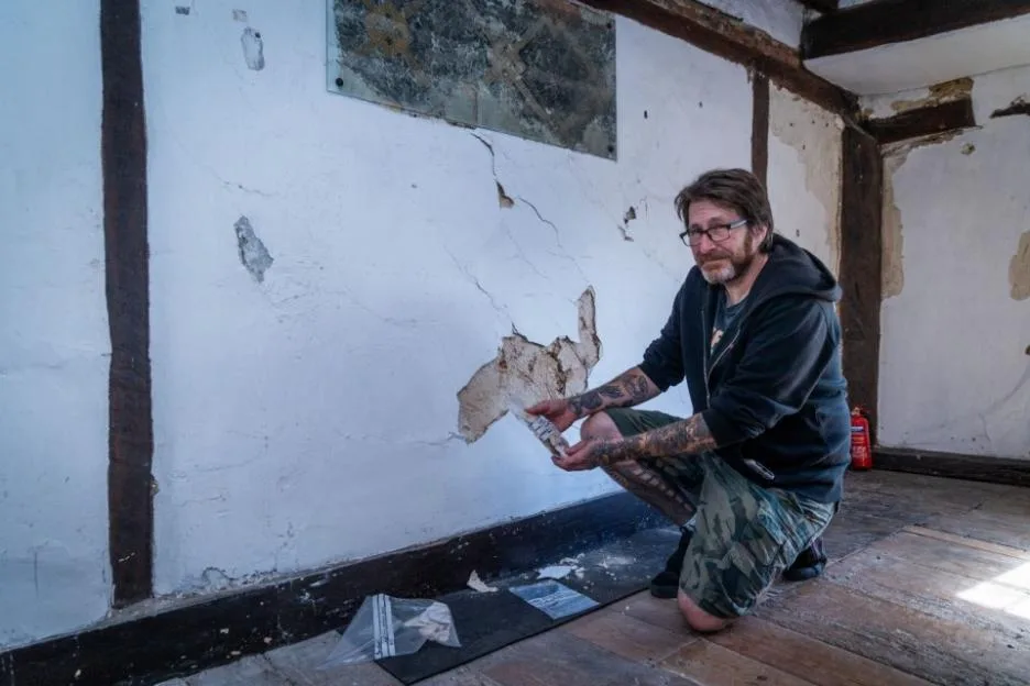 A tattooed man kneeling next to a damaged wall in an old building, holding a plastic bag containing plaster fragments.