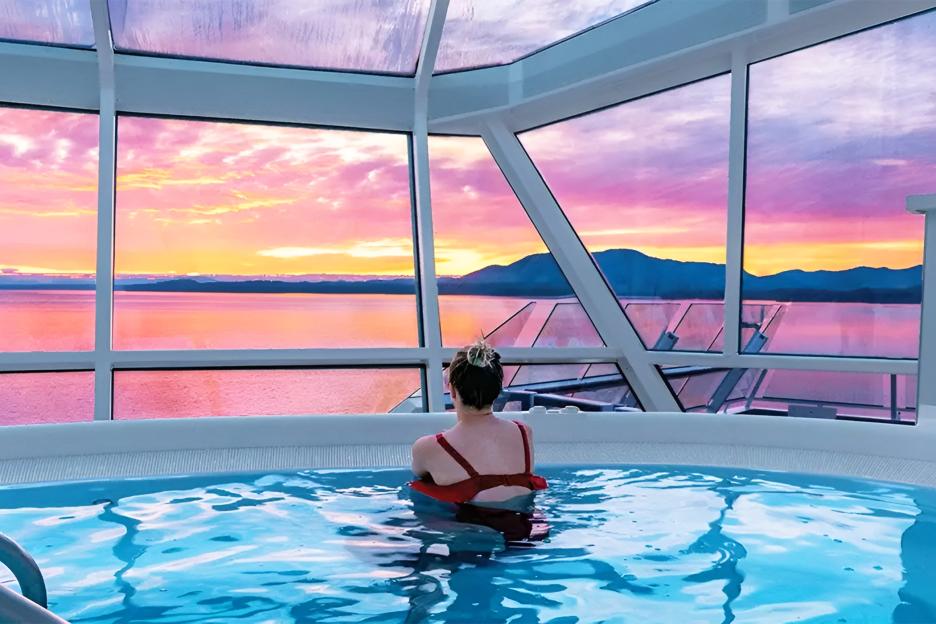 A woman in a hot tub facing large windows that overlook a body of water and mountains with a vibrant pink and orange sunset.