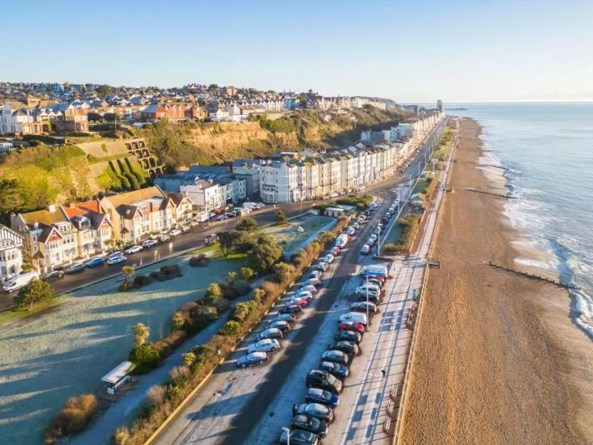 An aerial view of a coastal town with buildings along a clifftop and a road alongside a sandy beach.