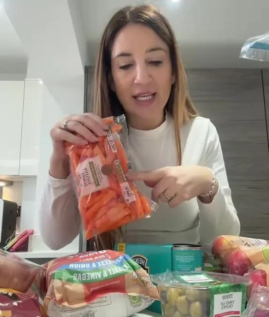 A woman points to a bag of baby carrots, part of a grocery haul including olives and chips.