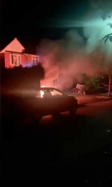 A dark image shows a car burning on a street at night, with smoke rising and a building in the background.