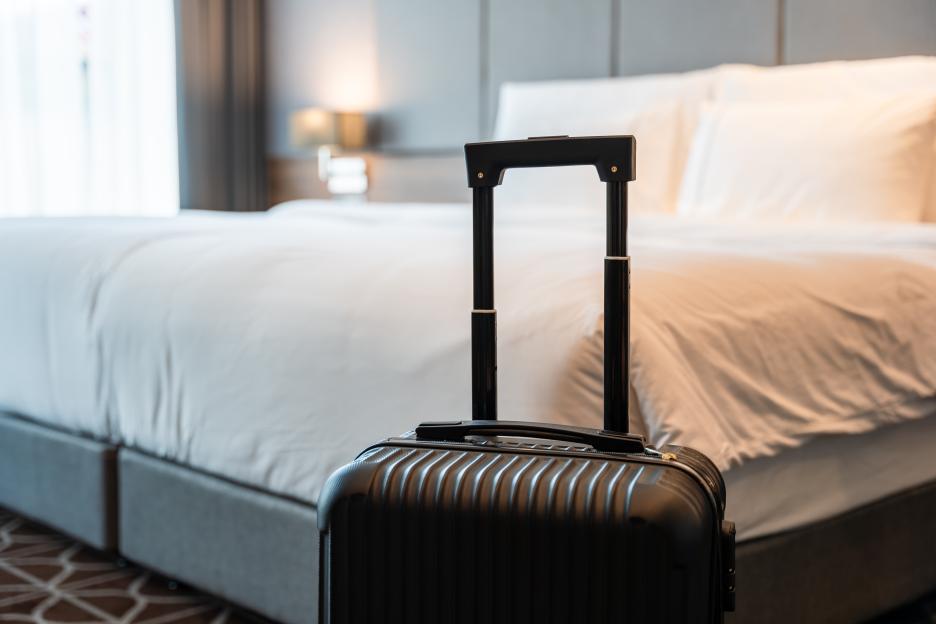 A black suitcase with its handle extended, sitting in a luxury hotel room in front of a white bed.