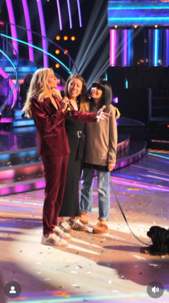 A woman in a red suit holding a microphone and talking to two other people and a dog on a stage with colorful lights.