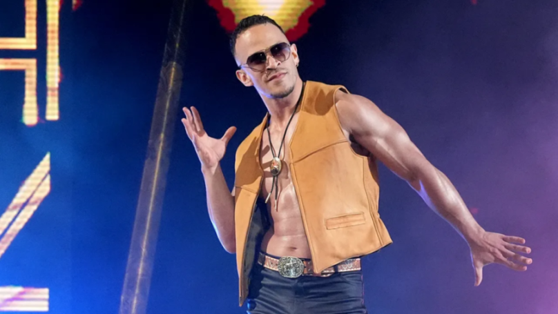 A man in a tan vest, black pants, sunglasses, and a bolo tie poses with his arms outstretched.