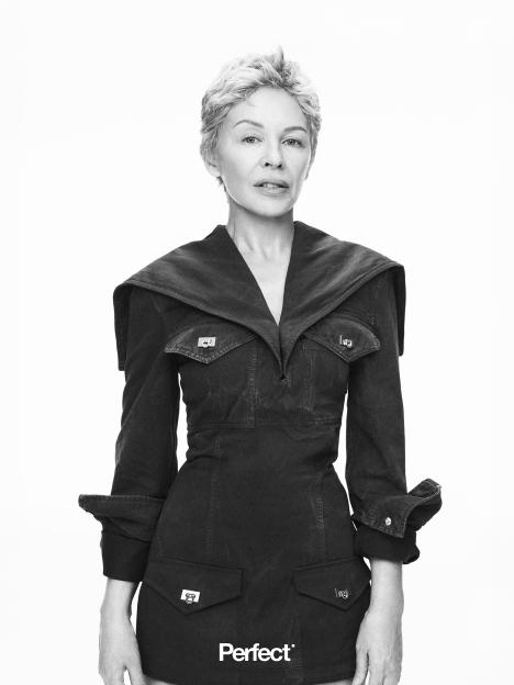 Black and white portrait of Kylie Minogue with short, grey hair, wearing a dark jacket with large lapels and prominent pockets, on a white background.