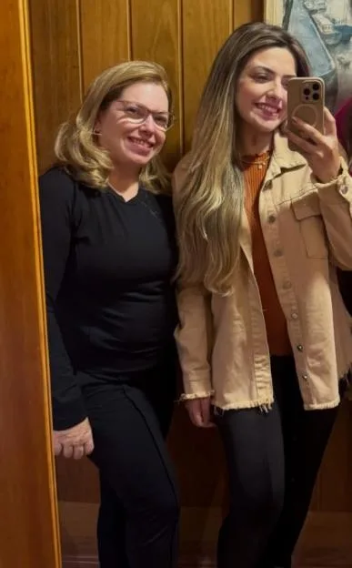 Two women pose in front of a wood-paneled wall, one with glasses and one holding a phone.