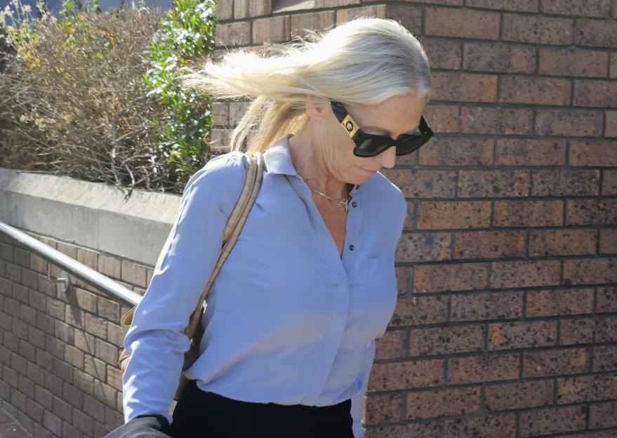 Sally-Anne Bowen in a light blue shirt and sunglasses walks past a brick wall.