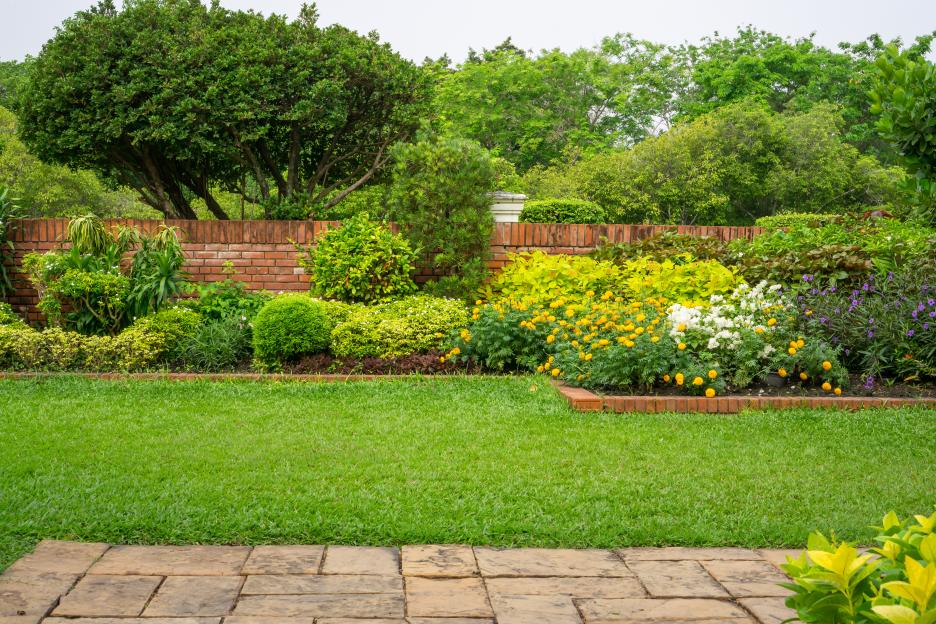 A vibrant backyard English cottage garden with colorful flowering plants, green grass, a brown pavement, and an orange brick wall.