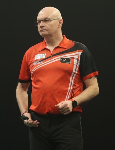 A darts player in an orange and black shirt and glasses looks to the right.