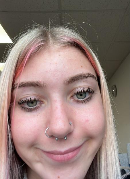 A smiling young woman with blonde and pink-streaked hair, green eyes, and nose piercings.