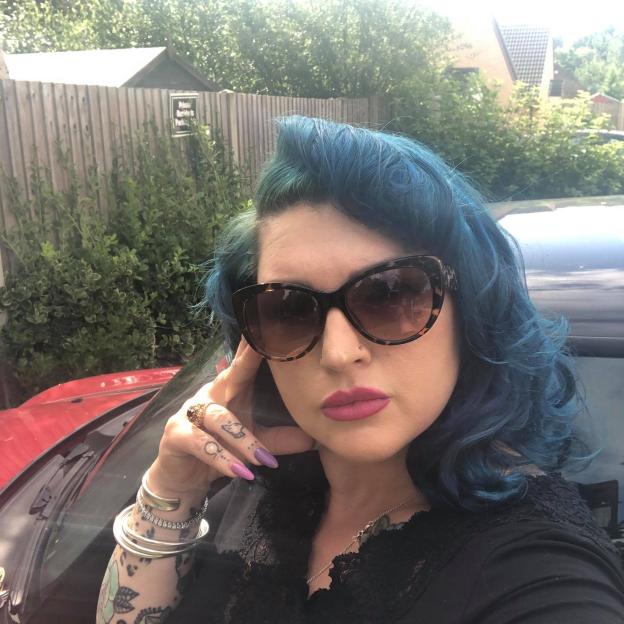 A woman with blue hair, sunglasses, and tattoos on her arm and fingers.