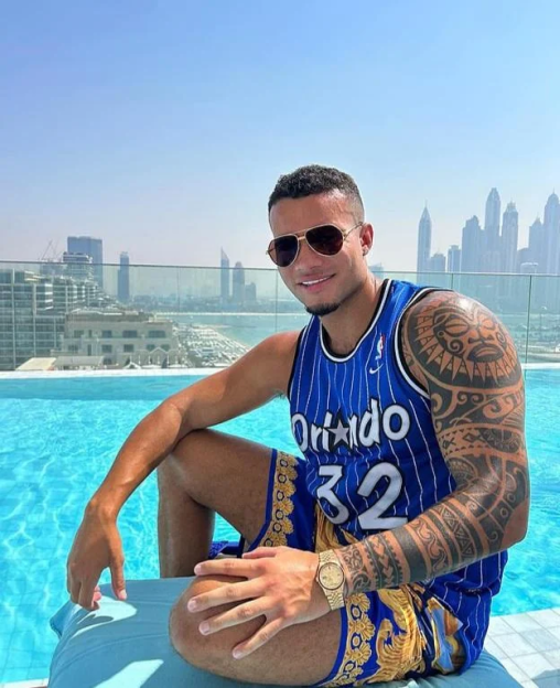 Harrison Sullivan, also known as HISTIKKYTOKKY, wearing an Orlando Magic jersey and shorts, sitting by a pool with a cityscape in the background.