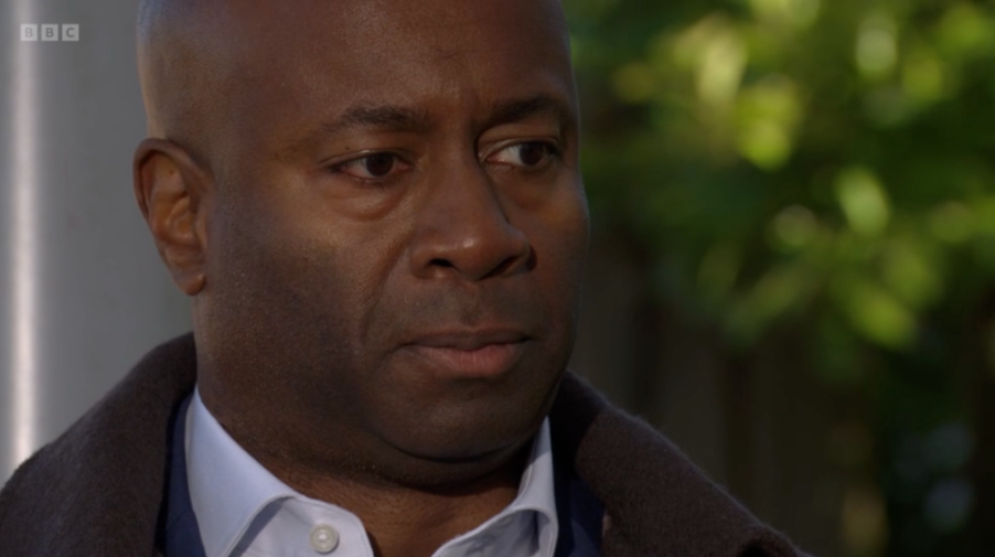 A man with dark skin and a bald head, wearing a blue shirt and brown jacket, looking intently to the right.