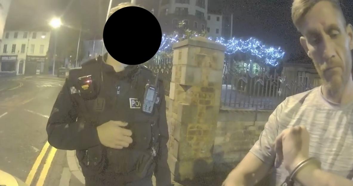 A police officer and a man, whose face is blurred, standing in front of him, at night. The man's wrist is handcuffed, and his face looks unhappy.