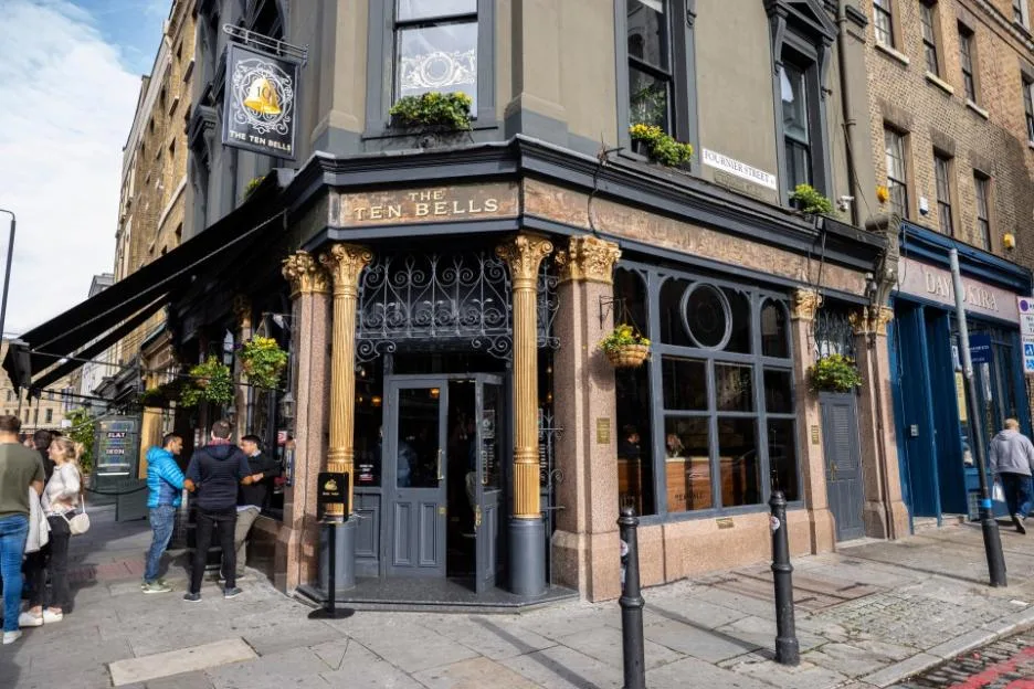 Exterior view of The Ten Bells pub in Spitalfields, London, featuring its ornate Victorian facade and a group of people standing outside the entrance.