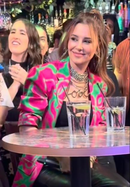 Cheryl Cole in a pink and green patterned blazer, layered necklaces, and a "Love" pendant.