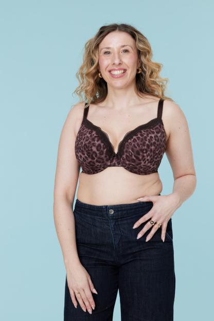 A woman with curly blonde hair and wearing a leopard print bra and dark jeans poses against a blue background.