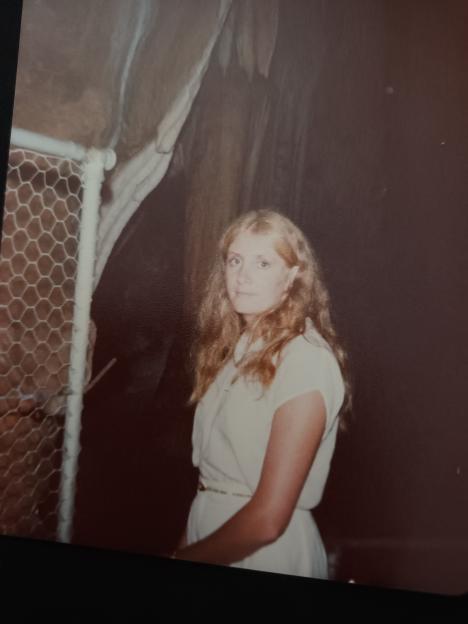 A young woman with long blonde hair and a white dress standing in a cave, looking directly at the camera.