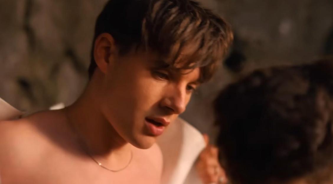 A shirtless man with brown hair and a silver necklace looks down and to his right, speaking to another person.