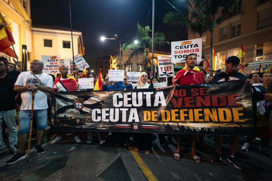 Residents of Ceuta protest at night, holding a large banner that reads "CEUTA NO SE VENDE, CEUTA SE DEFIENDE" (Ceuta is not for sale, Ceuta is defended).
