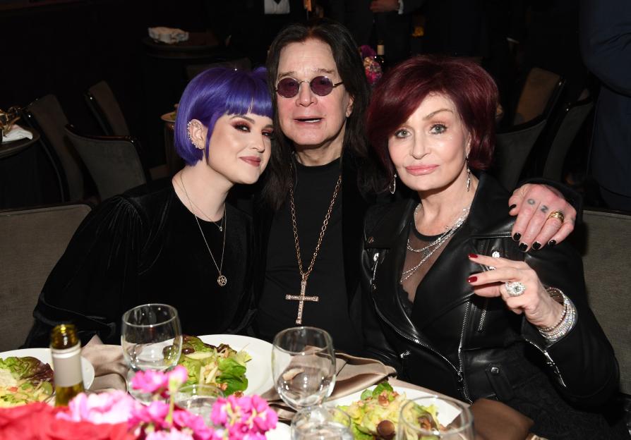 Kelly Osbourne, Ozzy Osbourne, and Sharon Osbourne sitting at a table with plates of salad and wine glasses.