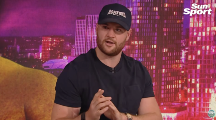 A man wearing a "ROMFORD BULL" cap and a dark t-shirt with his hands clasped, speaking in front of a digital city backdrop.