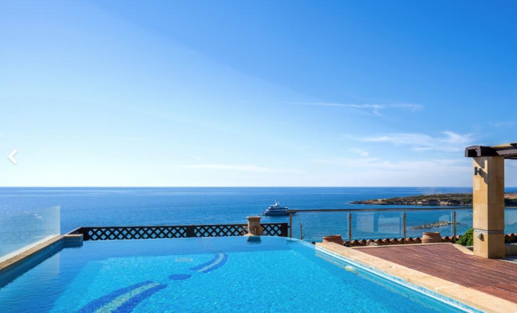 An infinity pool with blue tiles and a tiled deck overlooking the ocean with a yacht.