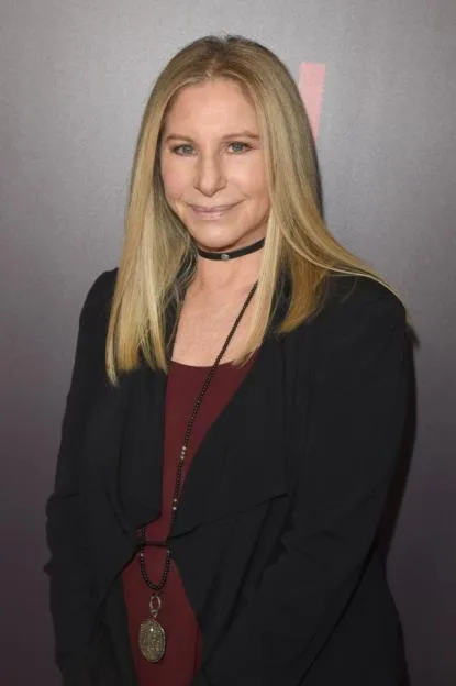 Barbra Streisand wearing a black blazer over a maroon top, with long blonde hair and a black choker and long beaded necklace, smiling at a Netflix event.